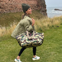 A woman walking through the highlands in Scotland wearing a large tote bag with an army print and sequin detailing in shades of gold, green, brown, and grey.