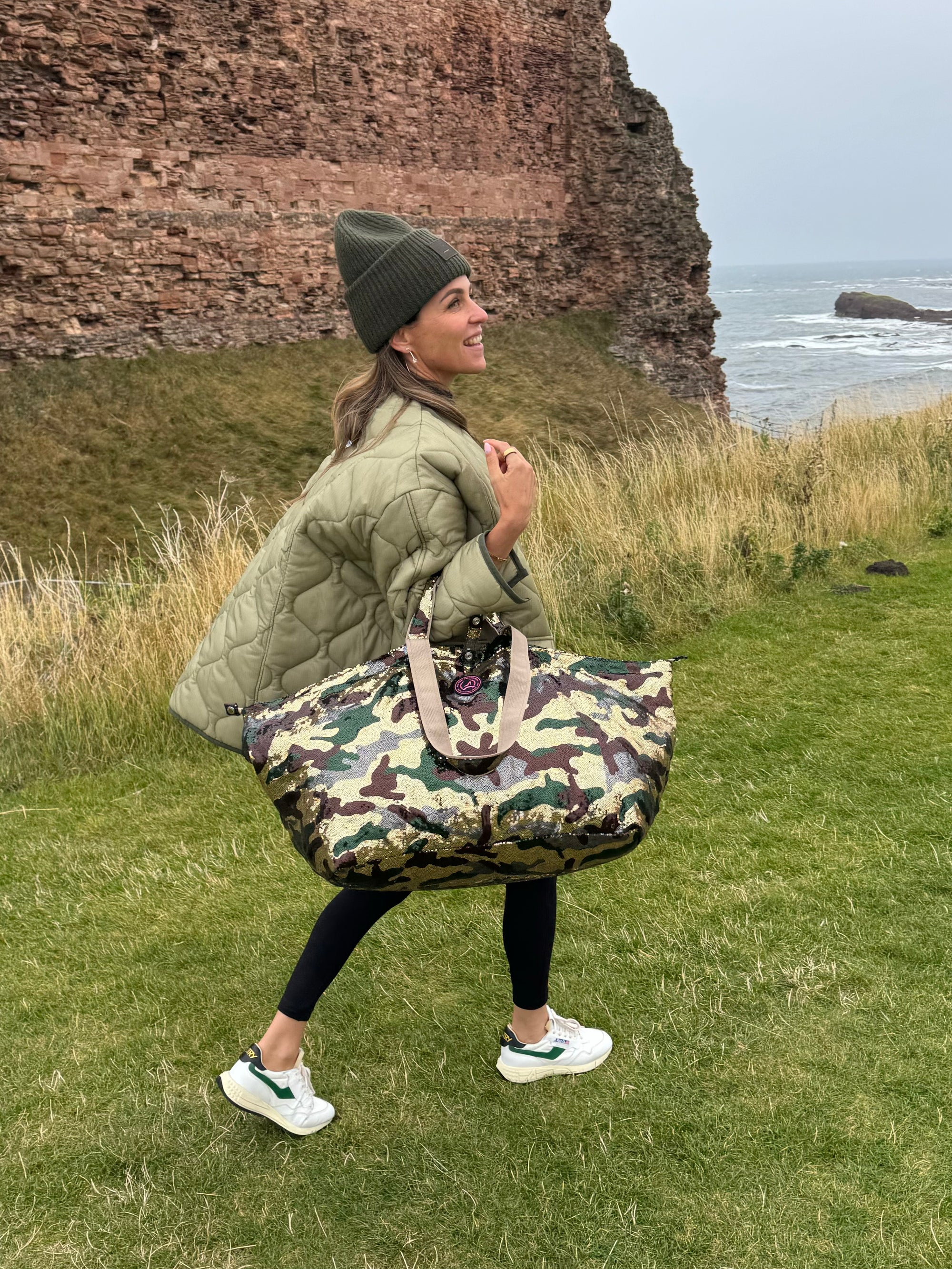 A woman walking through the highlands in Scotland wearing a large tote bag with an army print and sequin detailing in shades of gold, green, brown, and grey.