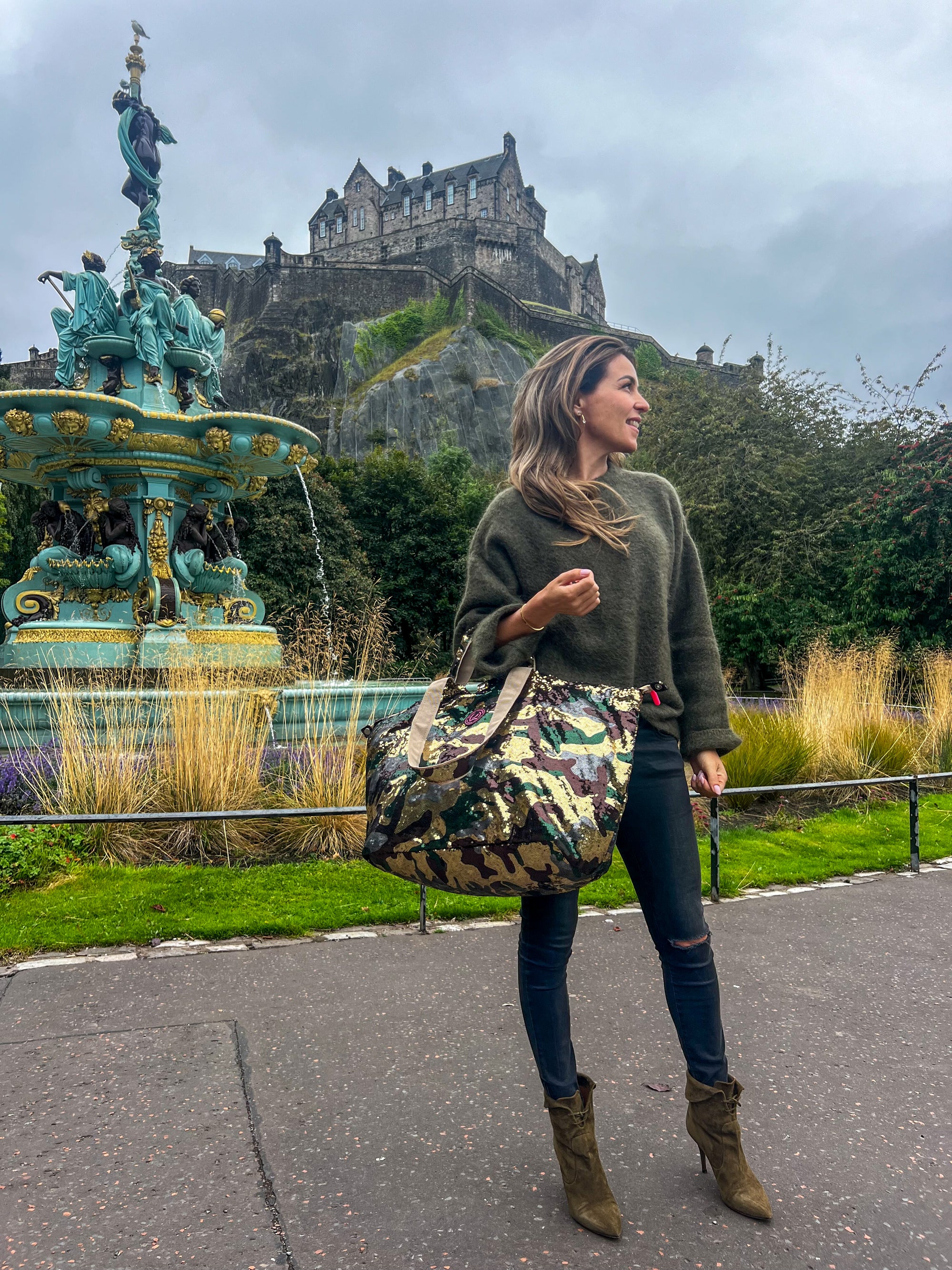 Woman standing in front of Edinburgh castle wearing a medium-sized tote bag with an army print and sequin detailing, predominantly in shades of green, gold, brown, and grey.