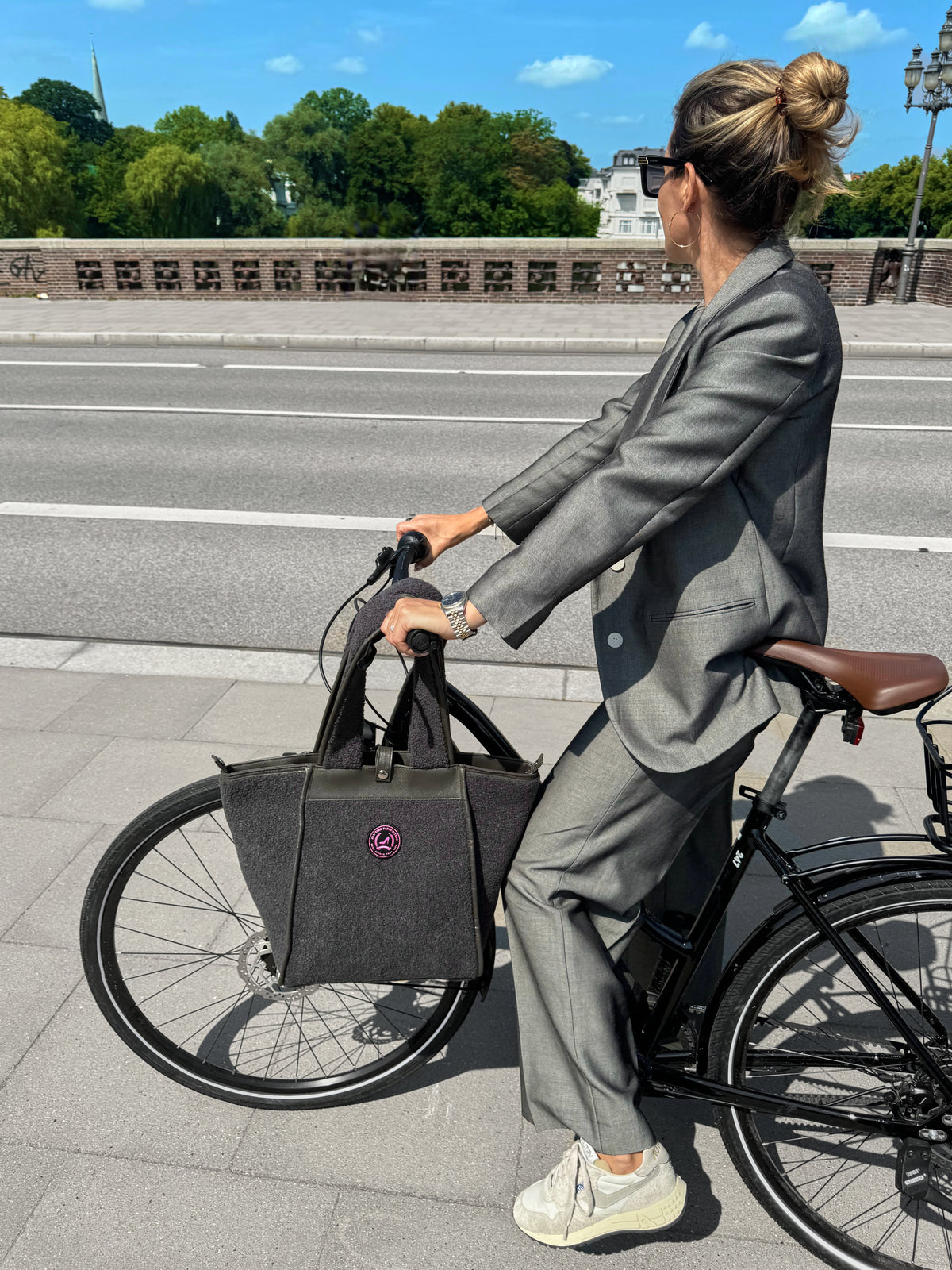 Vrouw in een grijs pak en sneakers op een zwarte fiets, met een grijze HAMBURG-draagtas van teddystof van All-time Favourites.