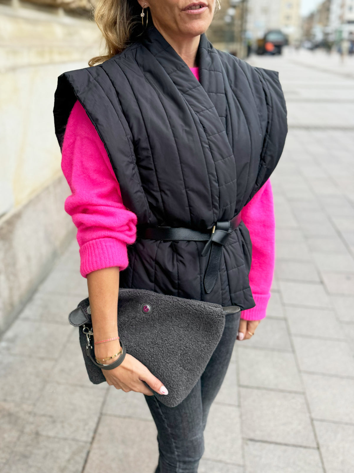 Woman holding grey teddy fabric toiletry bag HAMBURG on city sidewalk, wearing black vest and pink sweater.