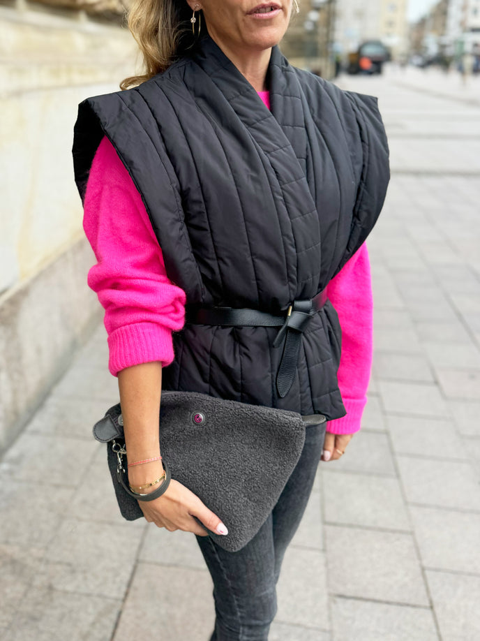 Vrouw die een grijze toilettas van teddystof met het opschrift HAMBURG vasthoudt op een trottoir in de stad, gekleed in een zwart vest en een roze trui.
