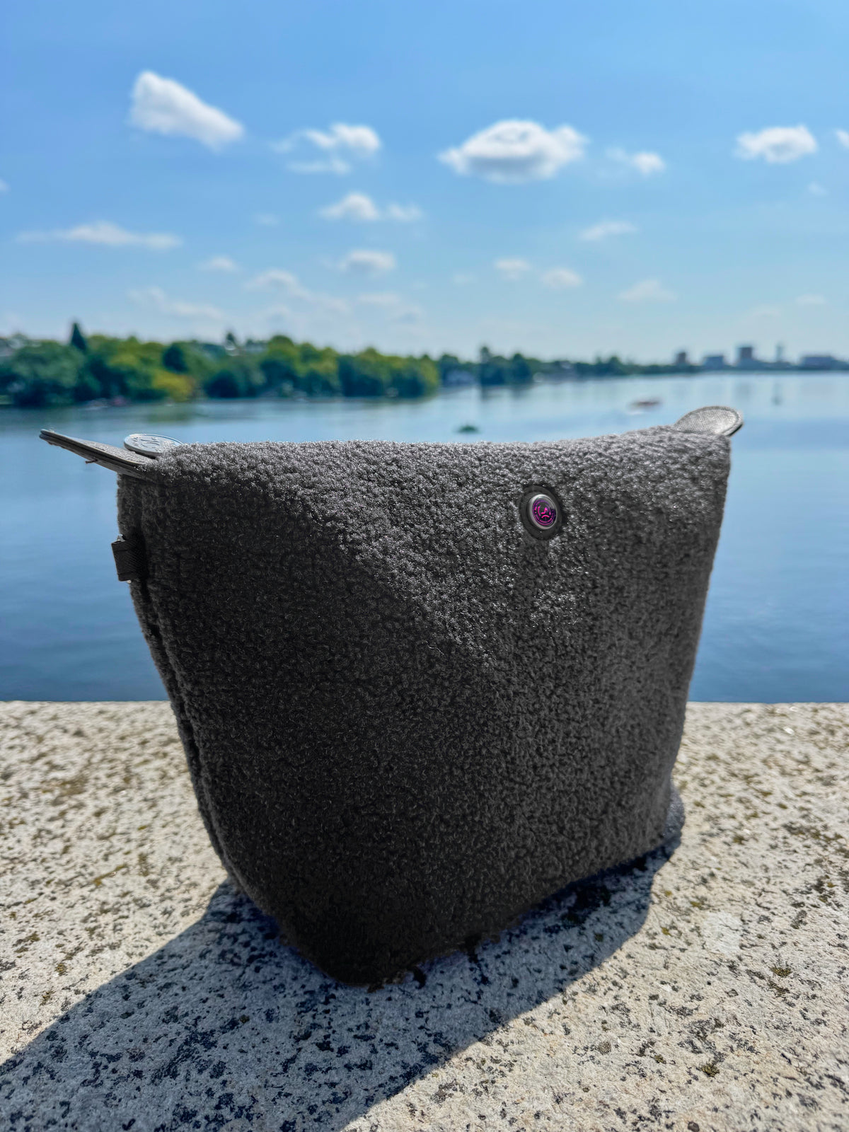 Grey teddy fabric toiletry bag from All-time Favourites on a stone ledge by a lake.