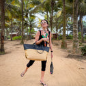 A woman in sunglasses, a colorful scarf, and black clothes walks on a sandy path lined with palm trees. She carries the Medium Tote Bag Black & Beige AUDREY from the beach resort collection and a small black bag while wearing flip-flops.