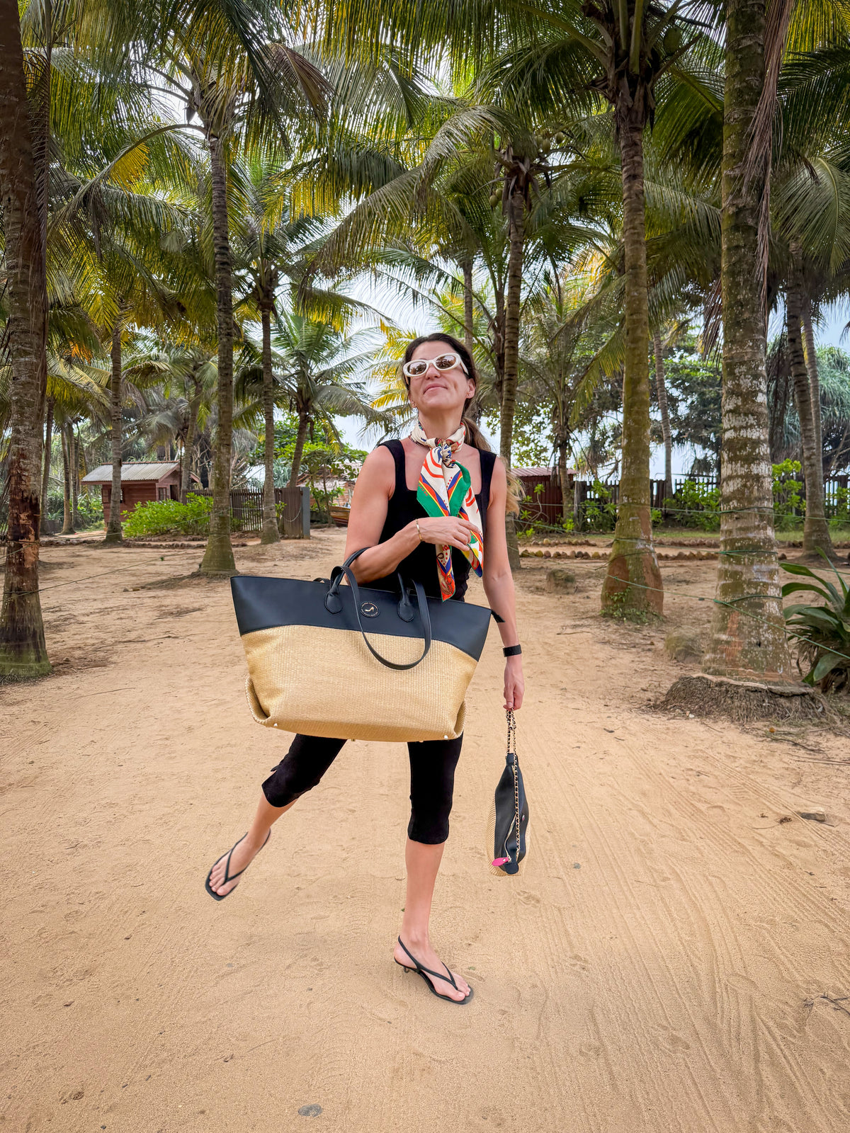 A woman in sunglasses, a colorful scarf, and black clothes walks on a sandy path lined with palm trees. She carries the Medium Tote Bag Black & Beige AUDREY from the beach resort collection and a small black bag while wearing flip-flops.