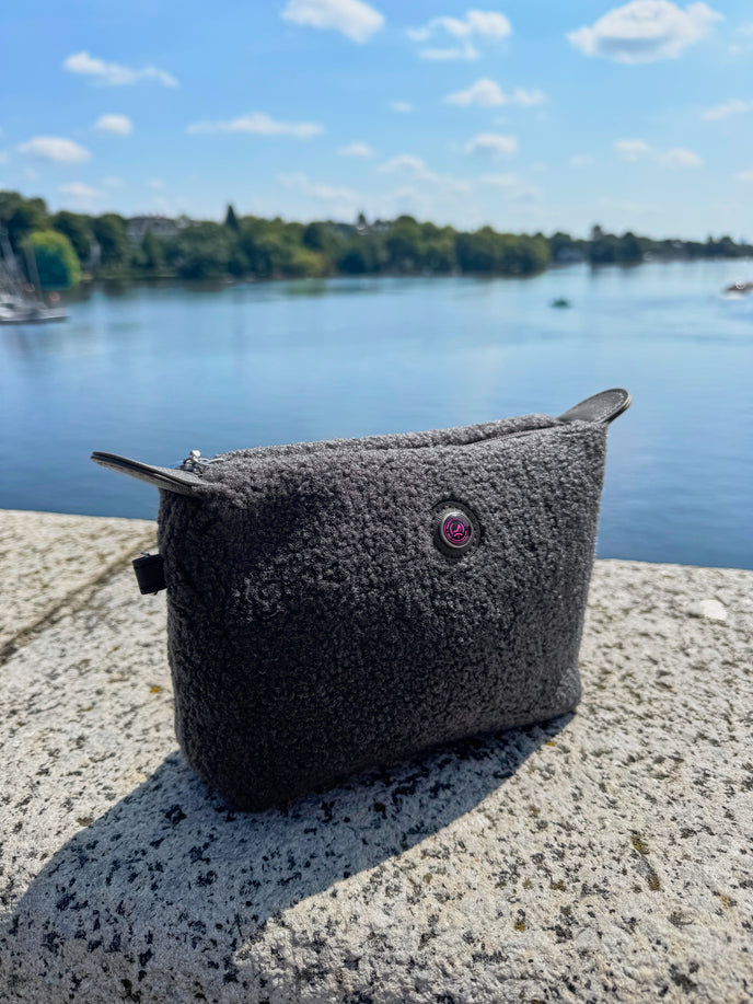 Grijze teddy-make-uptas HAMBURG op een stenen richel aan de oever van een meer, met een blauwe lucht en wolken, een luxe modeaccessoire.