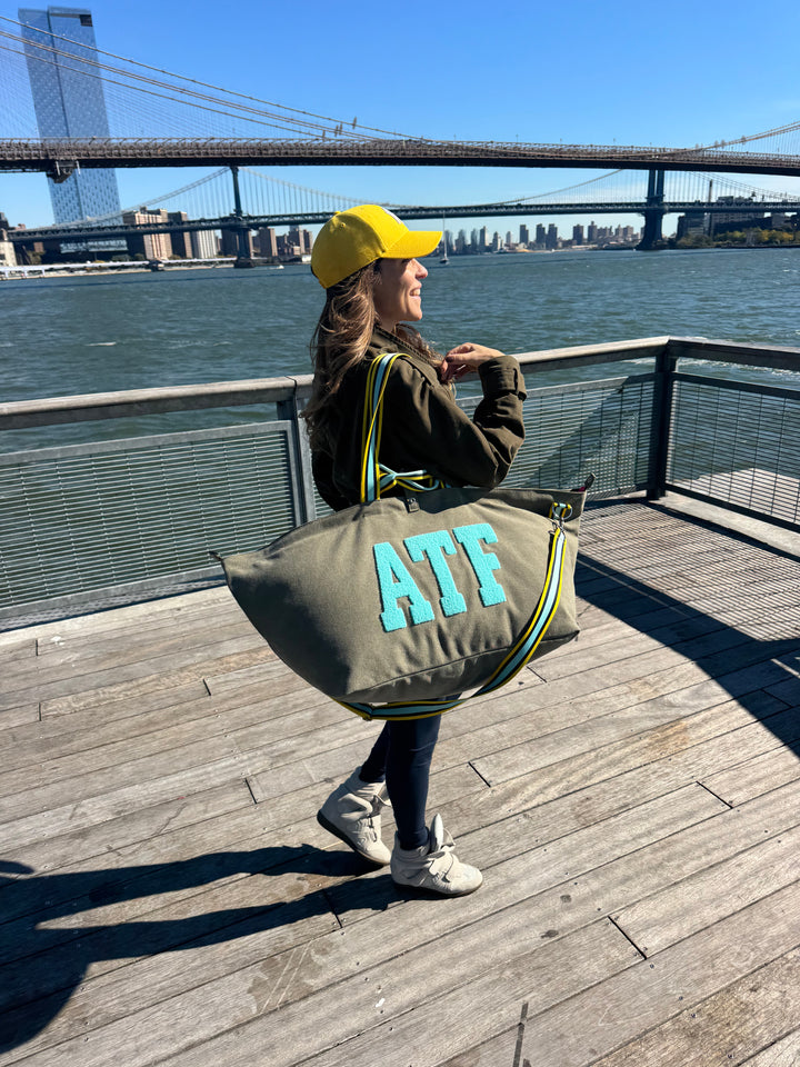 Vrouw met een gele pet die een grote BROOKLYN-draagtas van kaki canvas bij zich heeft op de promenade, met de stad en de brug op de achtergrond.