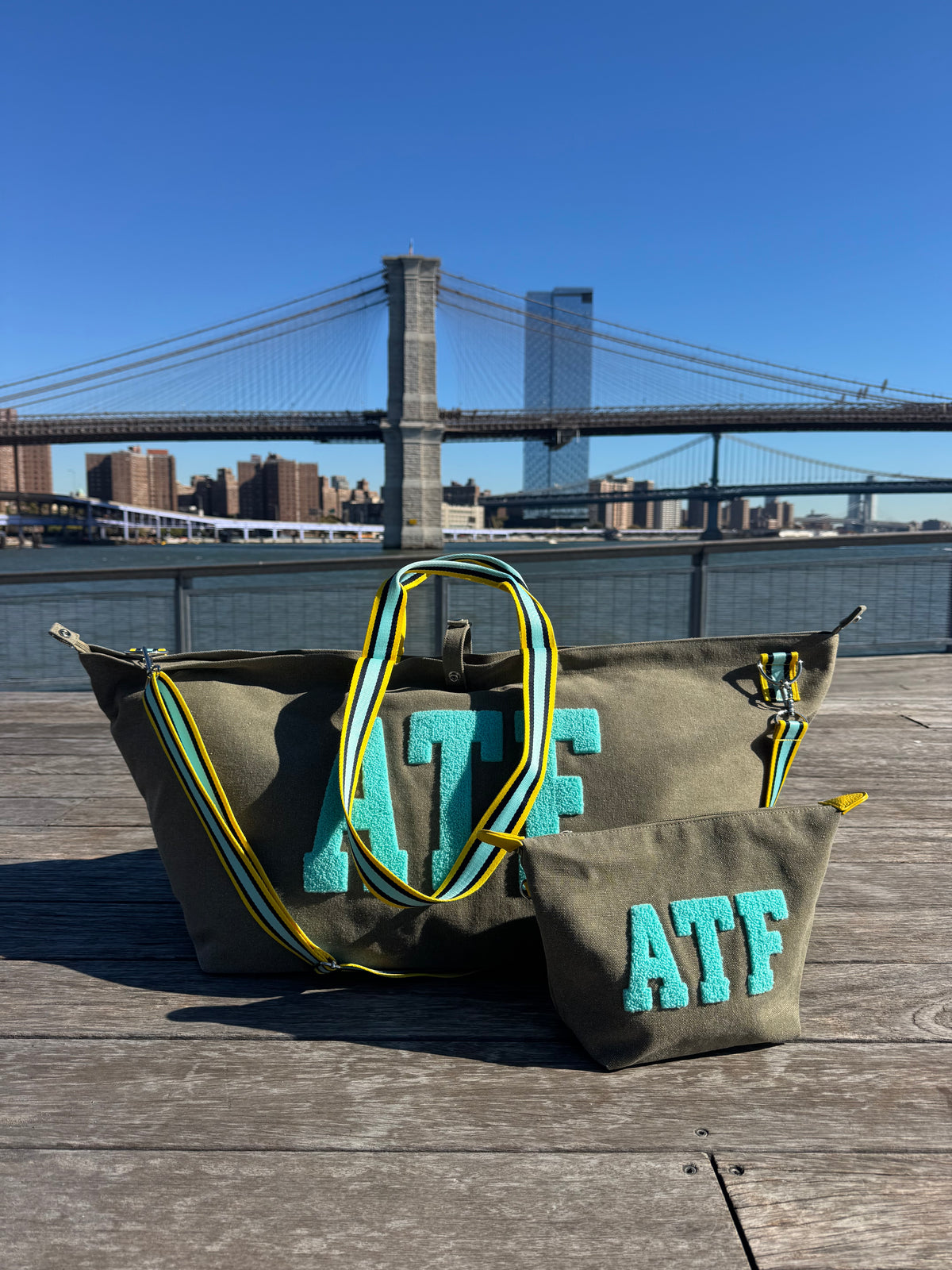 Two khaki BROOKLYN All-time Favourites large tote bags with yellow-blue straps on table, bridge in background.