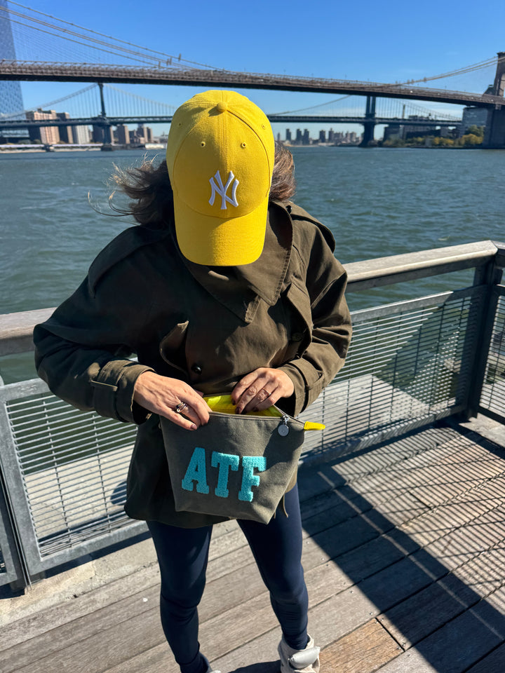 Iemand opent een kaki BROOKLYN-toilettas met een afbeelding van water en de skyline van de stad; hij draagt een gele NY-pet.