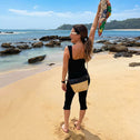 Woman walking on a sandy beach wearing a black crossbody bag and holding a colorful scarf