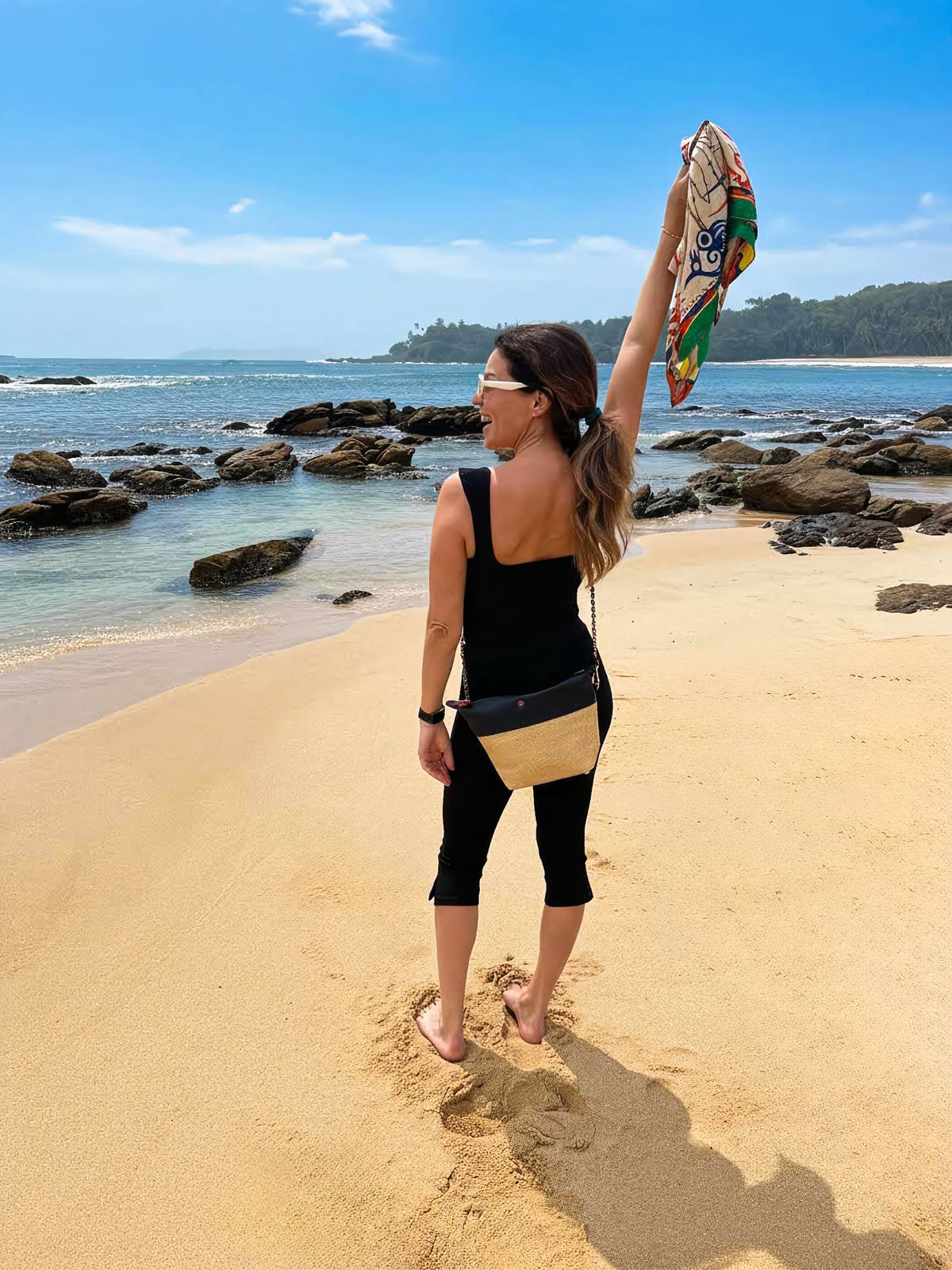 Woman walking on a sandy beach wearing a black crossbody bag and holding a colorful scarf