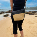 Woman wearing a black and beige straw crossbody bag on a sandy beach with rocks and ocean