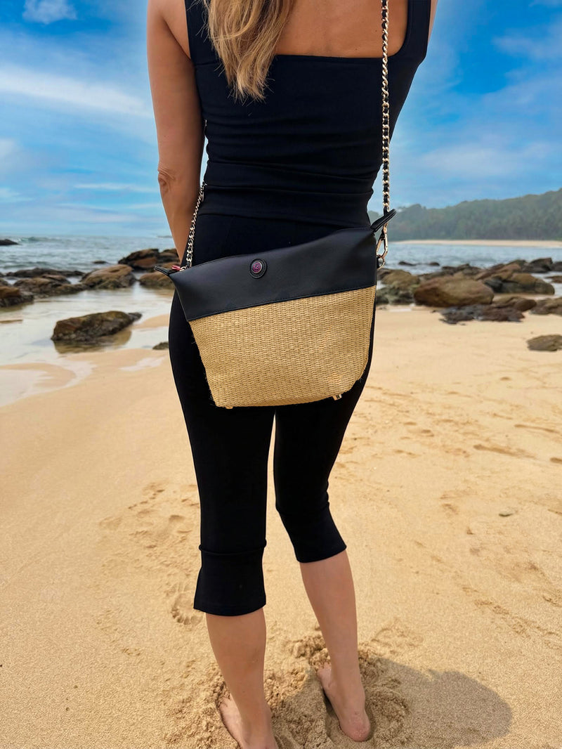 Woman wearing a black and beige straw crossbody bag on a sandy beach with rocks and ocean