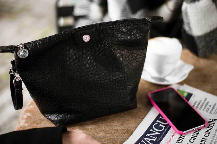 A black textured leather clutch bag with a wrist strap and silver zipper, resting on a wooden table next to a pink smartphone, a white coffee cup, and a newspaper.