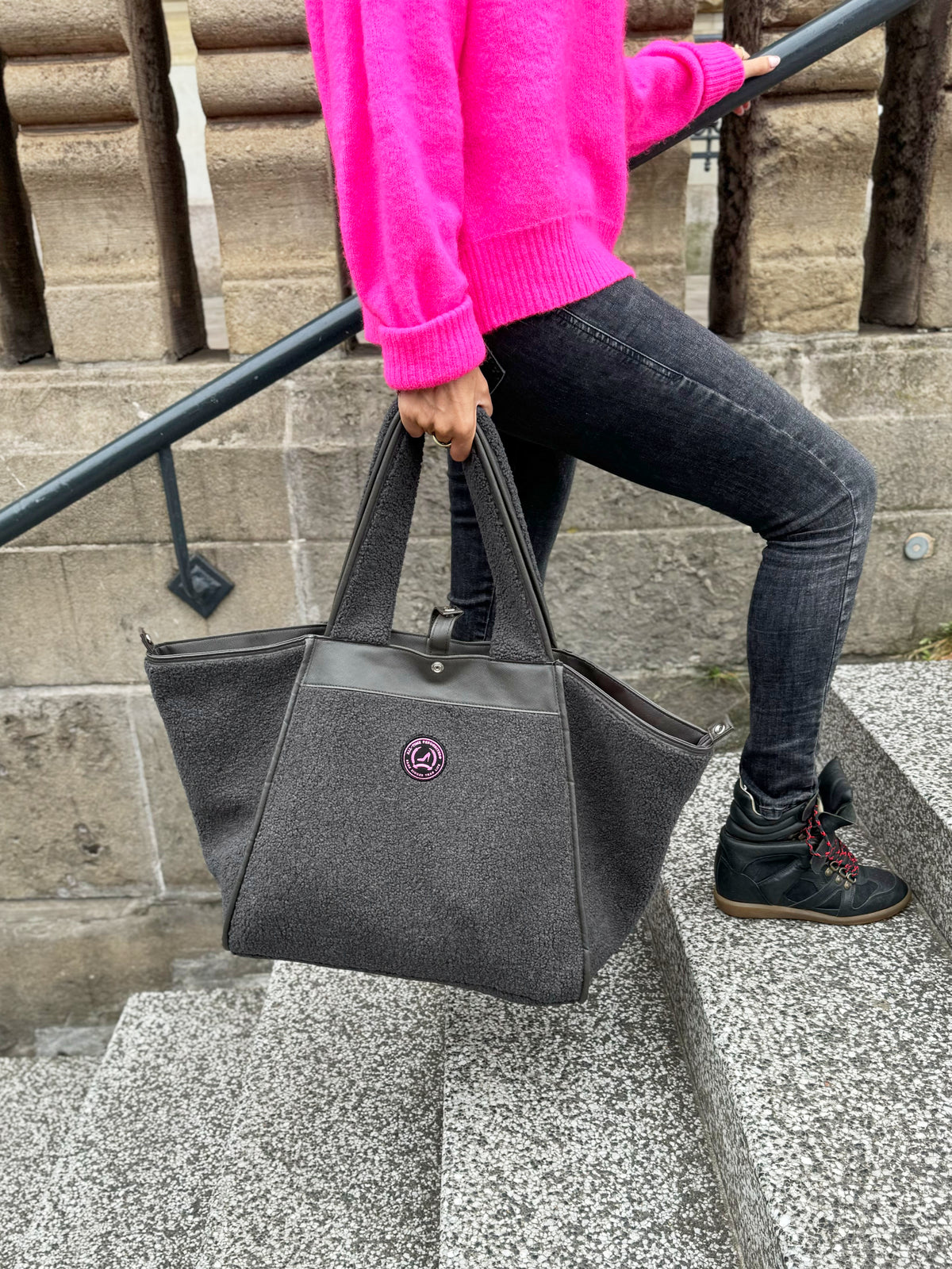 Eine Person in einem rosa Pullover trägt eine graue Teddy-Tasche in mittlerer Größe im HAMBURG-Stil die Steintreppe im Freien hinauf.