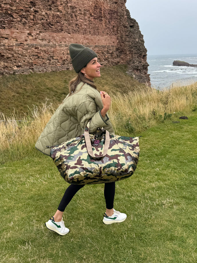 Woman in green jacket holding army print sequin tote bag by ocean cliffs.