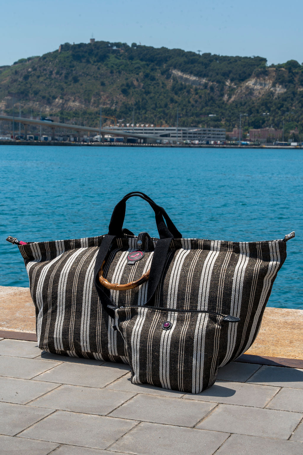 Striped All-time Favourites bag with bamboo handle on a stone surface by a body of water with a mountainous background