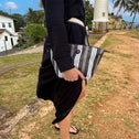 A woman in a black dress and sandals stands on grass outdoors, holding the Toiletry Bag Black & White GALLE. Behind her are a white lighthouse, palm trees, and buildings under a partly cloudy sky.
