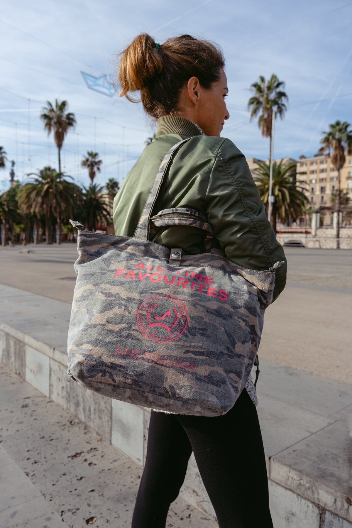 Vrouw in een groene jas met een kleine camouflage-draagtas bij zich, in de buurt van palmbomen, buiten.