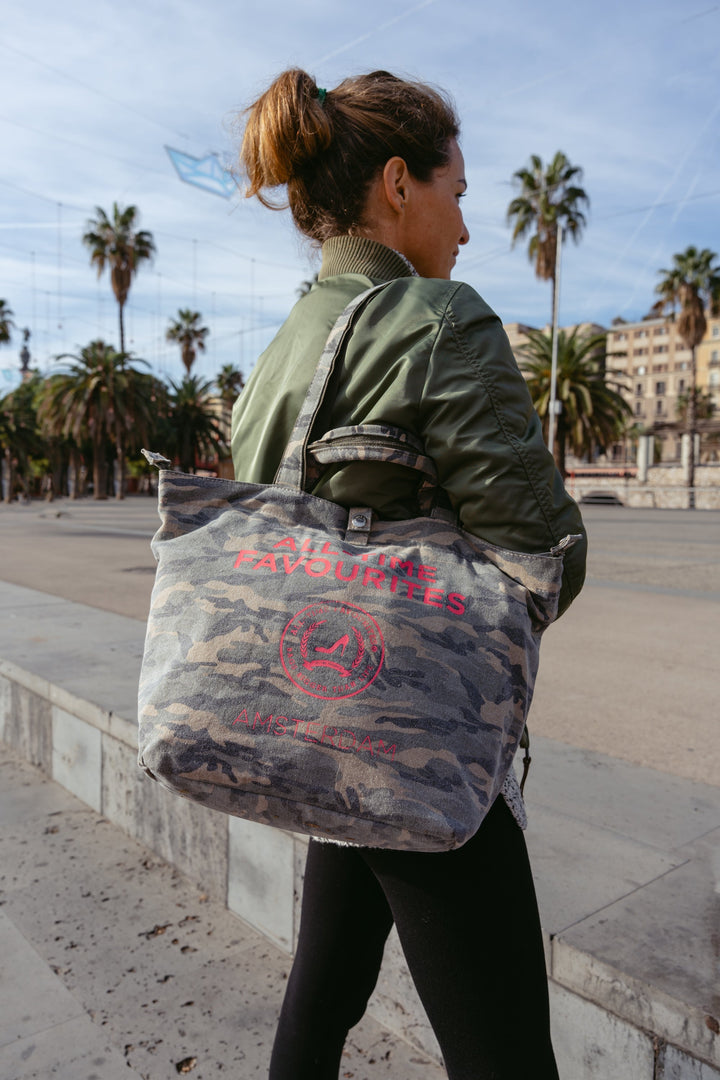 Frau in grüner Jacke, die eine kleine Tasche im Camouflage-Muster trägt, in der Nähe von Palmen, im Freien.