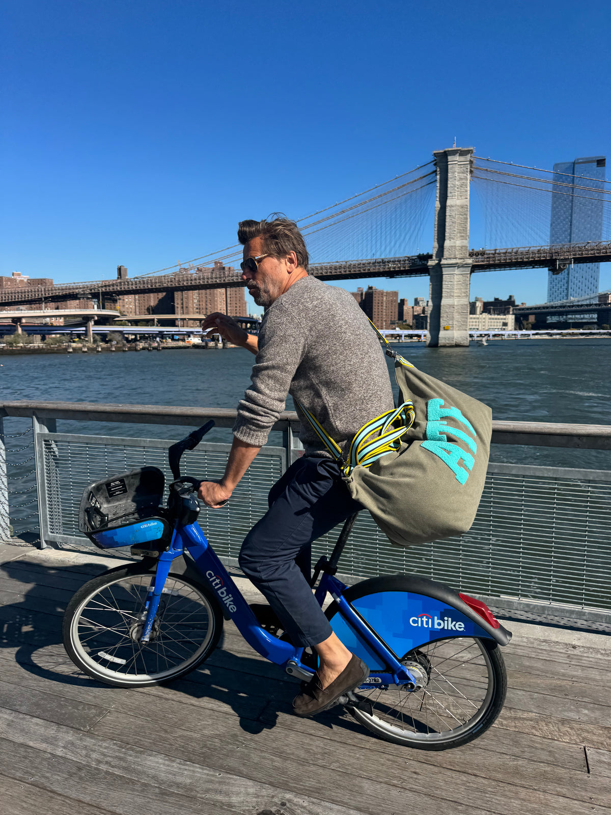 Man cycling by waterfront carrying khaki canvas large tote bag; Brooklyn city skyline in background.