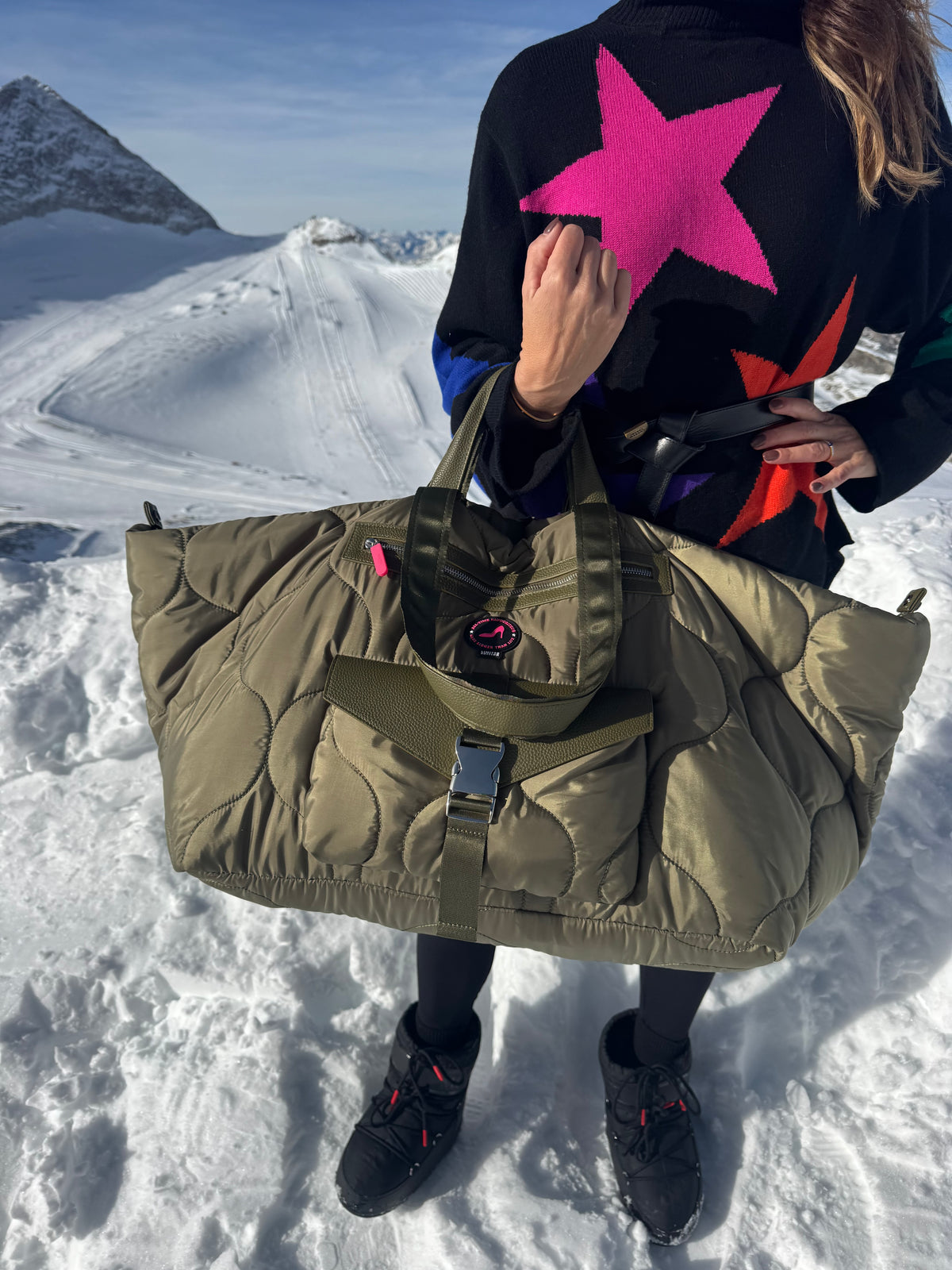 Person in snow holding puffy green DIANA Large Tote Bag with star sweater, ski resort mountains in background.