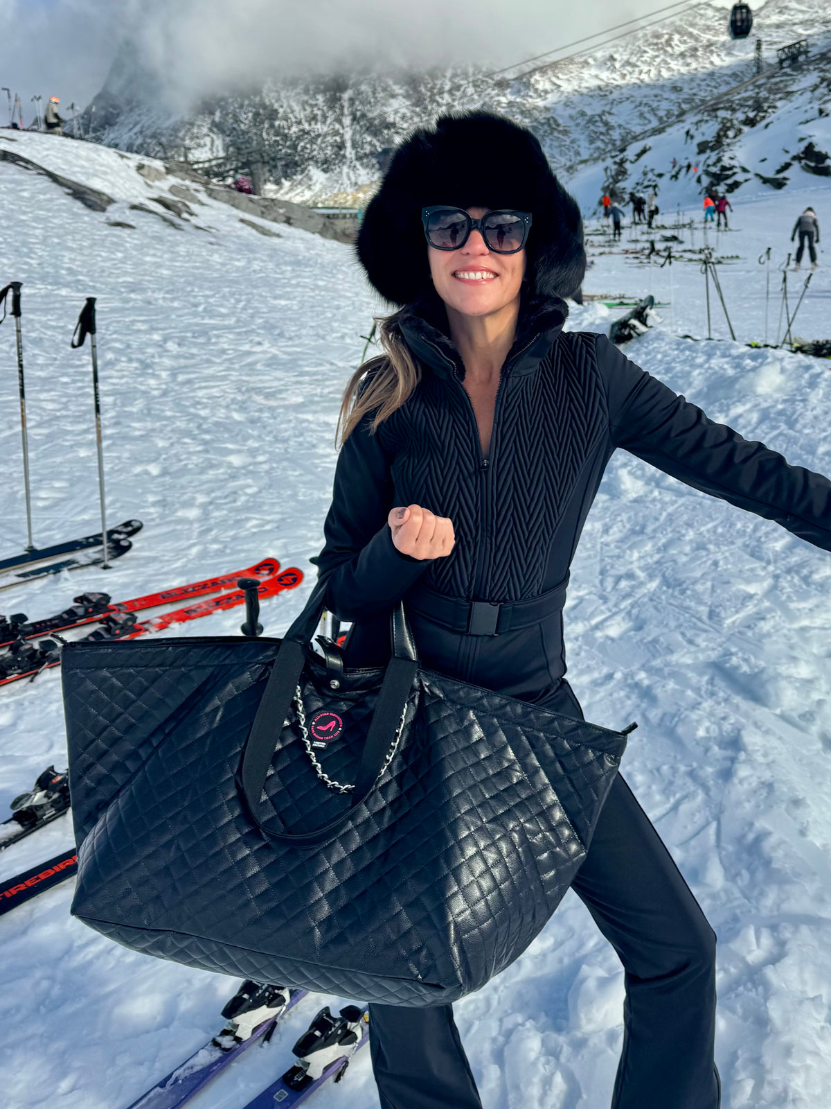 Eine Frau in schwarzer Winterkleidung und mit Pelzmütze steht auf einem verschneiten Hang und hält eine schwarze KATE Caviar Tote aus Leder sowie ihre Skier in der Hand.