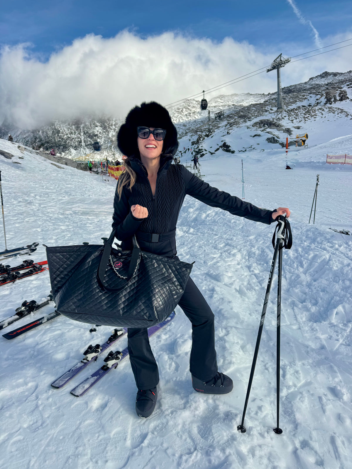 Eine Frau in schwarzer Winterkleidung, die Skier und eine große schwarze Ledertasche in der Hand hält, auf einem verschneiten Berg.