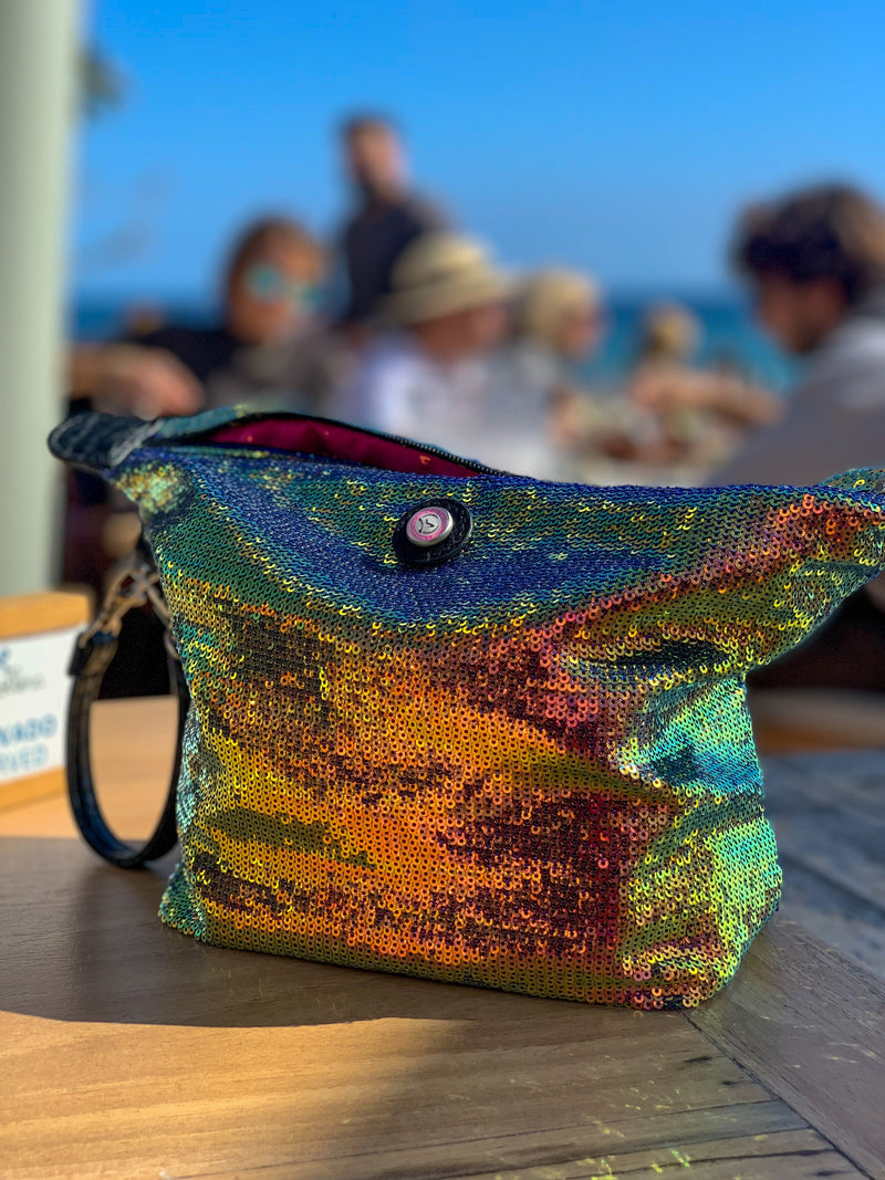Multicolored sequin toiletry bag with zipper on a wooden table, beach setting in the background
