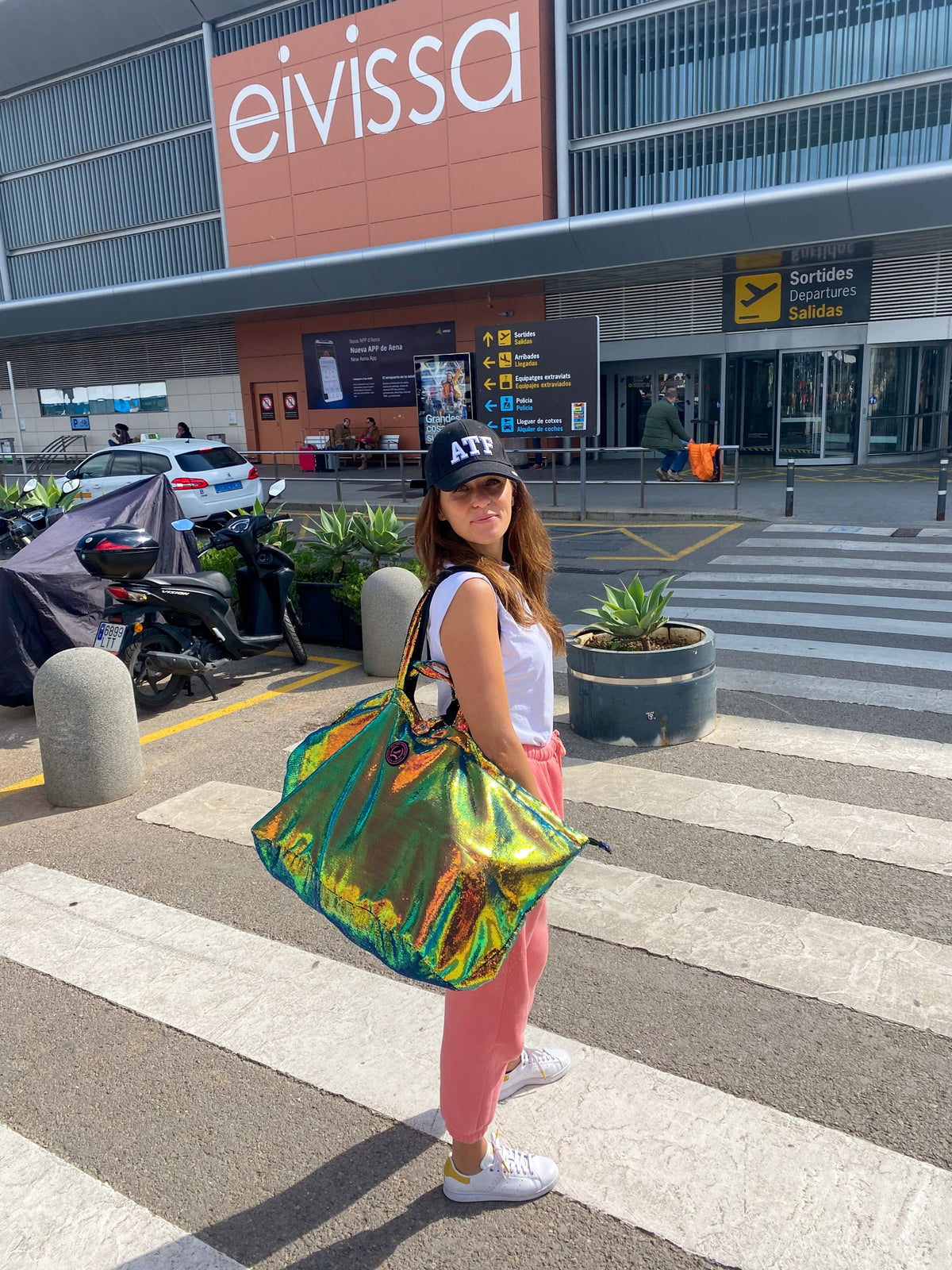 Een vrouw in een roze broek met een grote, veelkleurige tas met pailletten op de luchthaven van Eivissa.