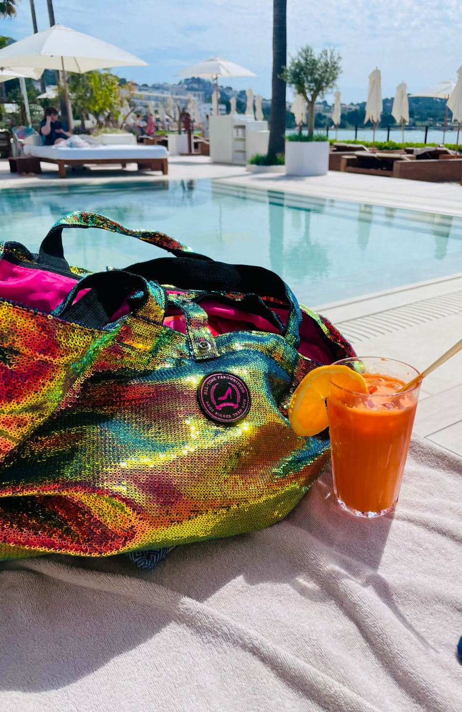 Veelkleurige tas met pailletten van All-time Favourites aan het zwembad, met een oranje drankje en parasolletjes.