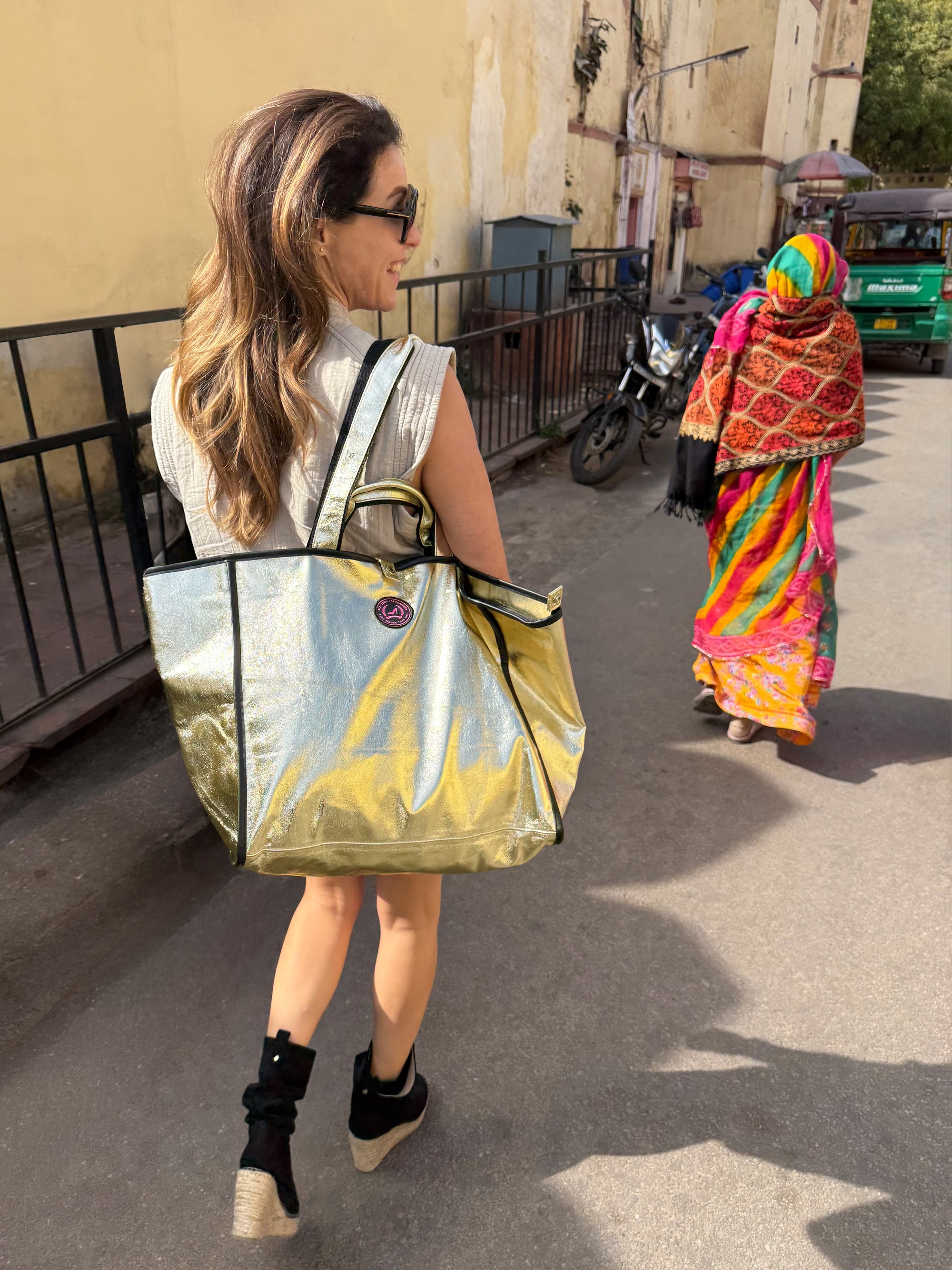 Woman with a reflective gold tote bag walking on a street in India with a colorful saree-wearing woman in the background.
