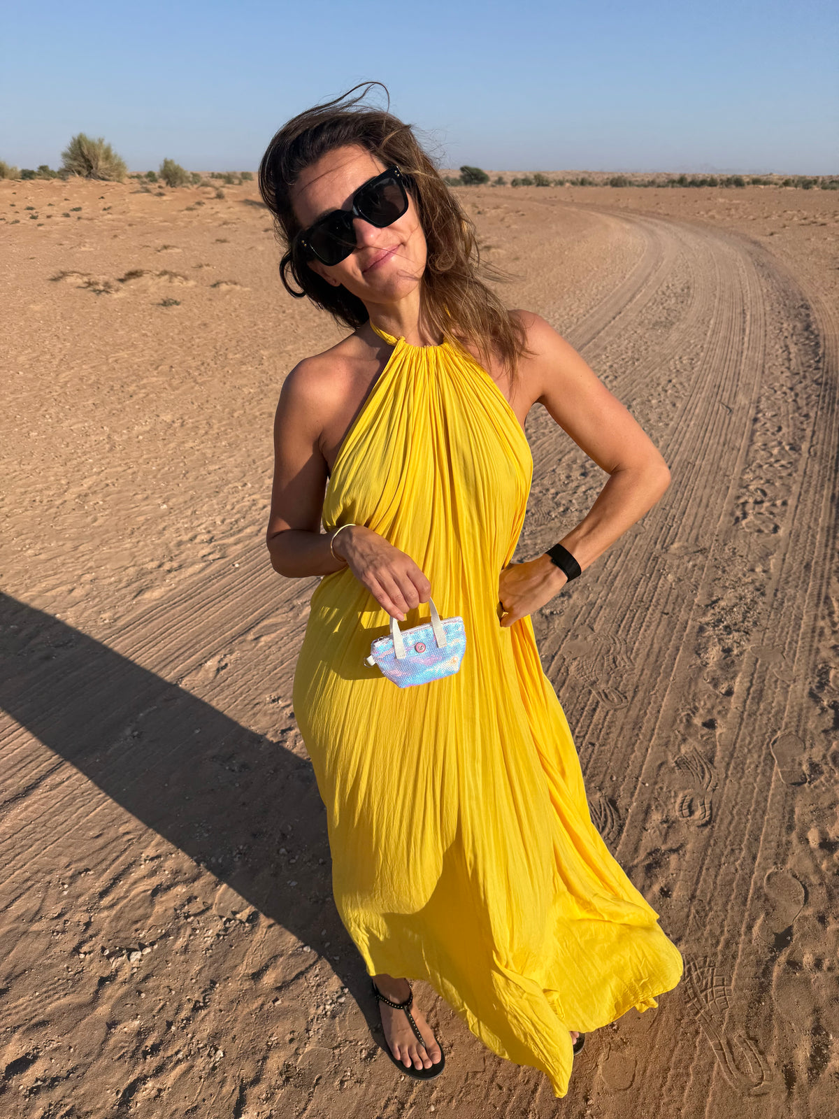 Frau in gelbem Kleid auf dem Sand, die eine weiße, mit Perlenpailletten besetzte Schlüsselhülle in der Hand hält und eine Sonnenbrille trägt.
