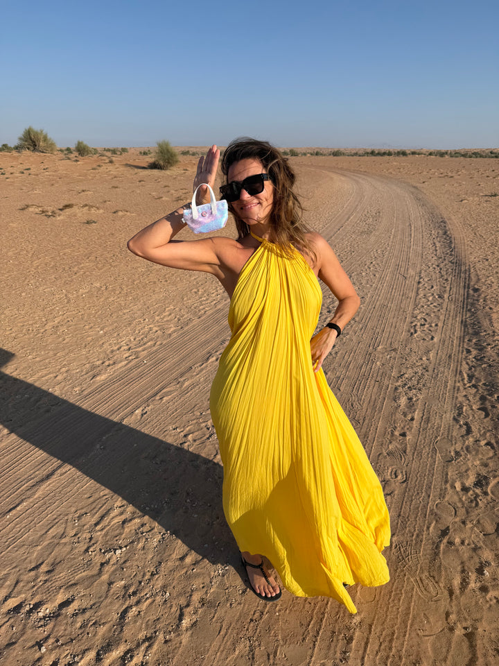 Vrouw in een gele jurk op het strand, met een wit tasje met parelmoeren pailletten en een zonnebril van All-time Favourites.