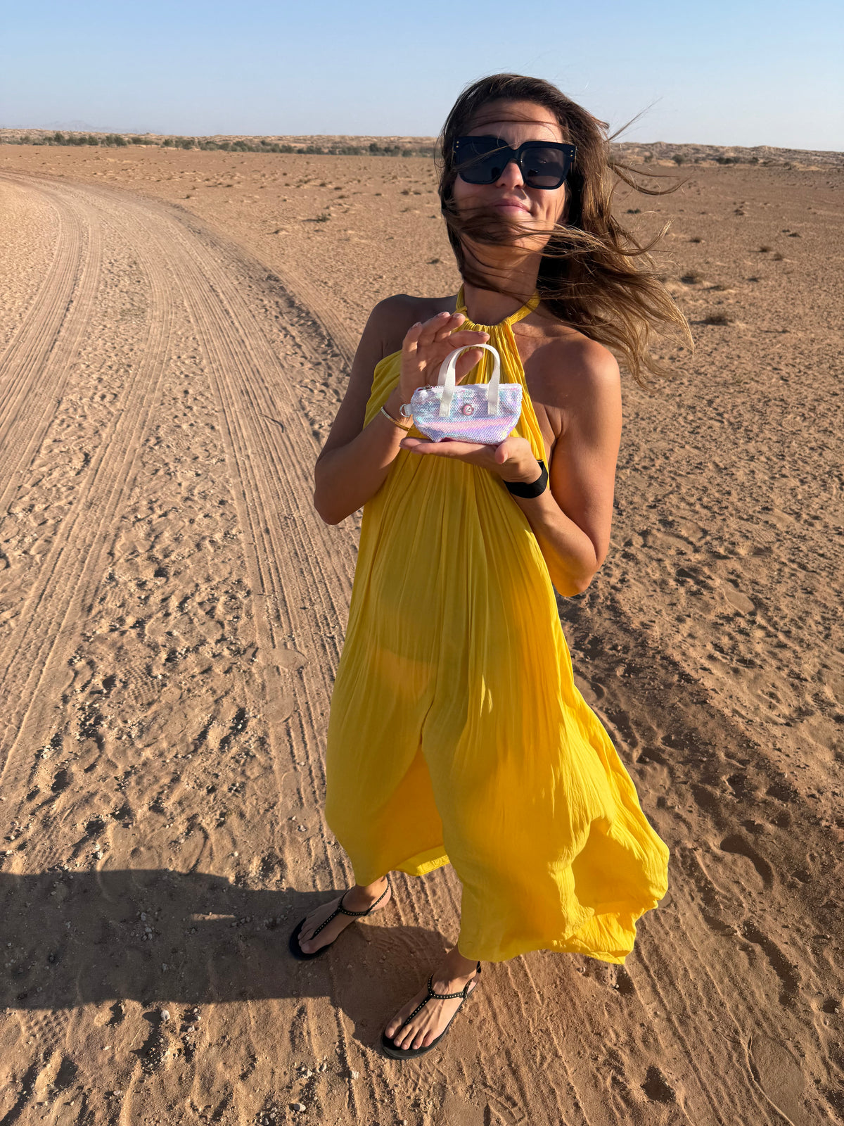 Eine Frau in einem gelben Kleid, die eine weiße, mit Perlenpailletten besetzte Schlüsseltasche in der Hand hält, auf einem Wüstenweg.