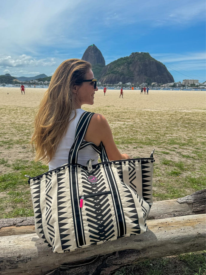 Rio Medium Tote Bag carried over the shoulder, featuring black pom-pom trim, front zip pocket and geometric black and off-white pattern, photographed on the beach.
