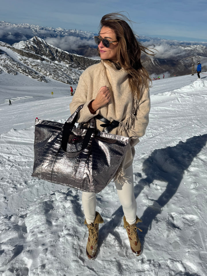 Woman in beige outfit holds BRIGITTE bronze leather tote bag on snowy mountain, wearing boots and sunglasses.