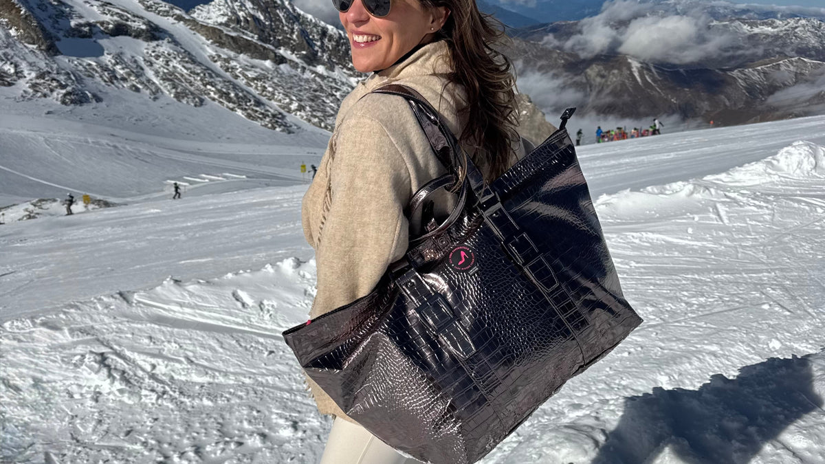 Eine Frau mit Sonnenbrille, die eine mittelgroße, bronzefarbene Metallic-Tragetasche der Marke BRIGITTE auf einem verschneiten Berghang in der Hand hält.