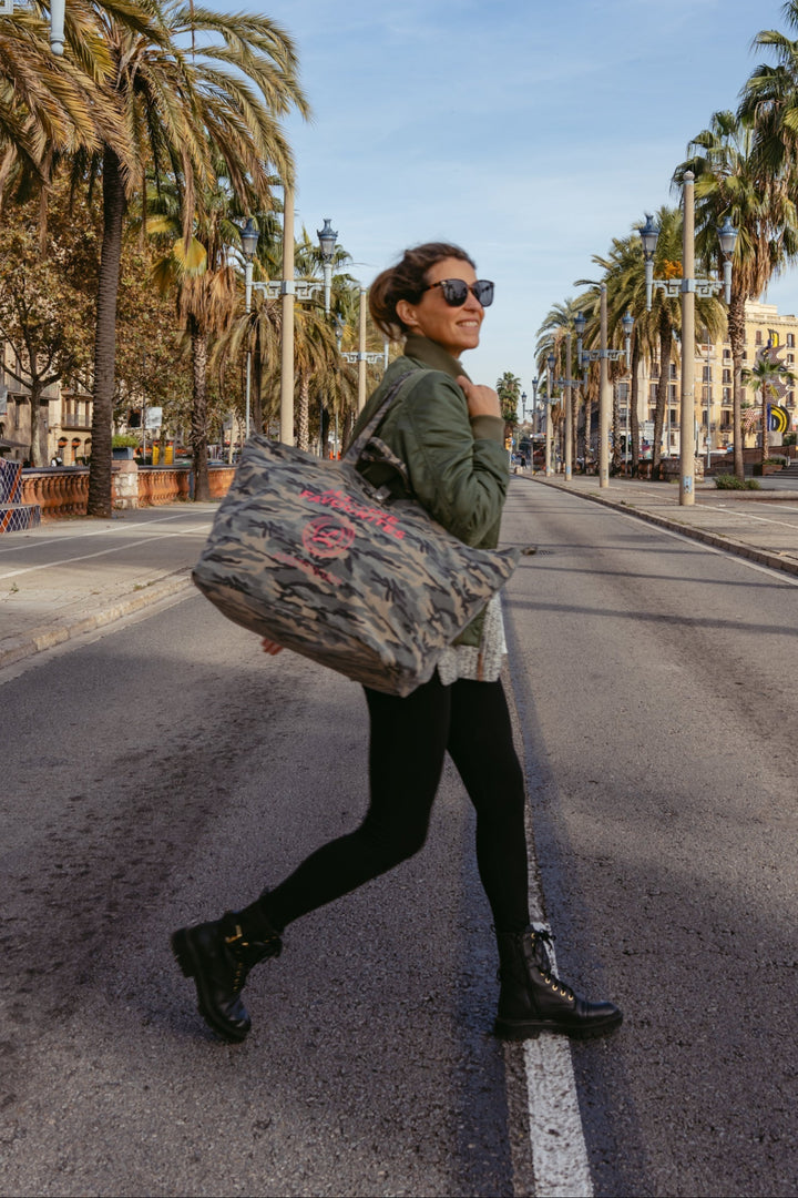 Eine Frau mit Sonnenbrille trägt eine mittelgroße „All-time Favourites“-Tragetasche im Camouflage-Muster auf einer von Palmen gesäumten Straße.