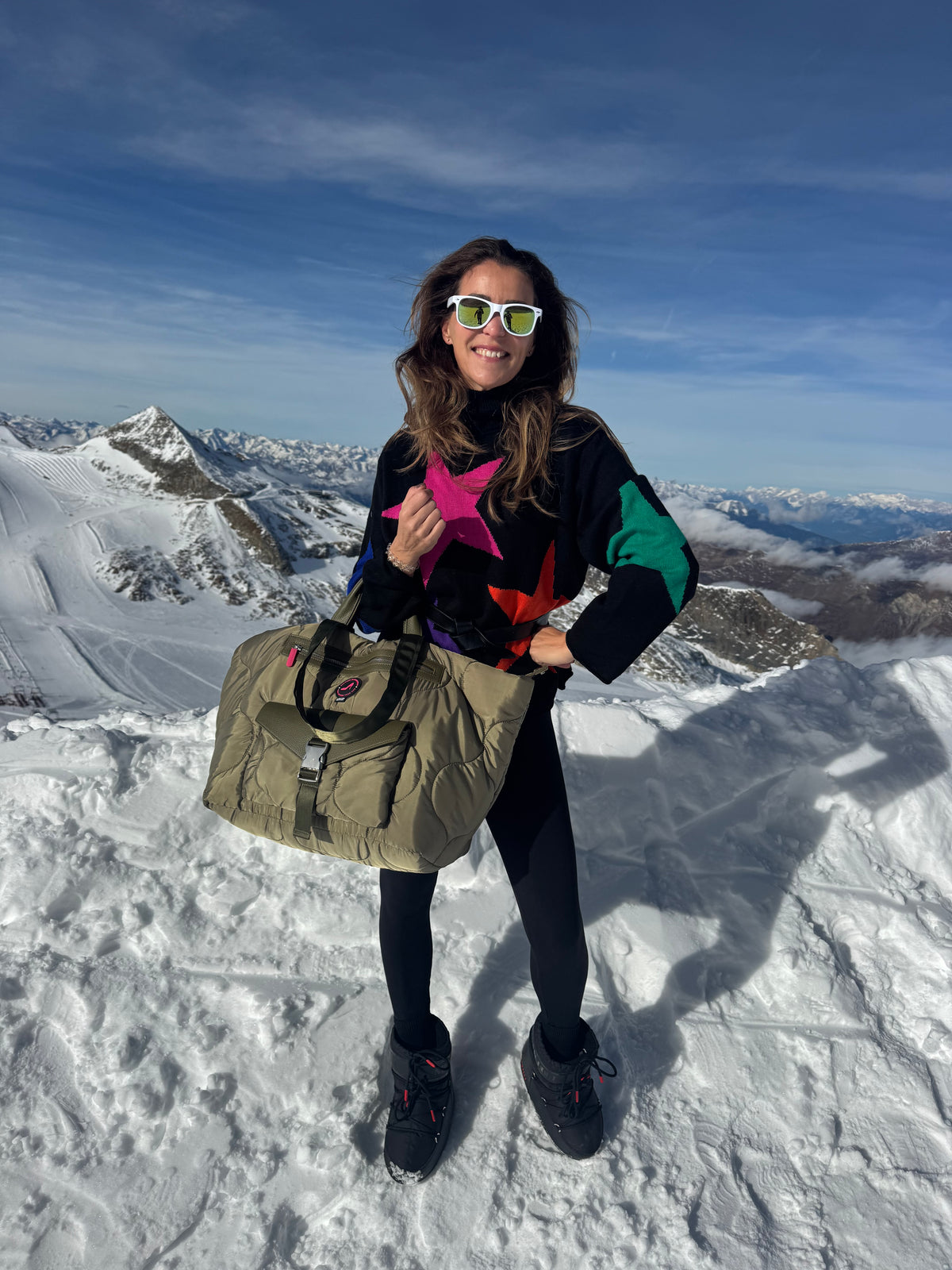 Woman with sunglasses and sweater holds green puffy medium tote bag DIANA on snowy mountain.