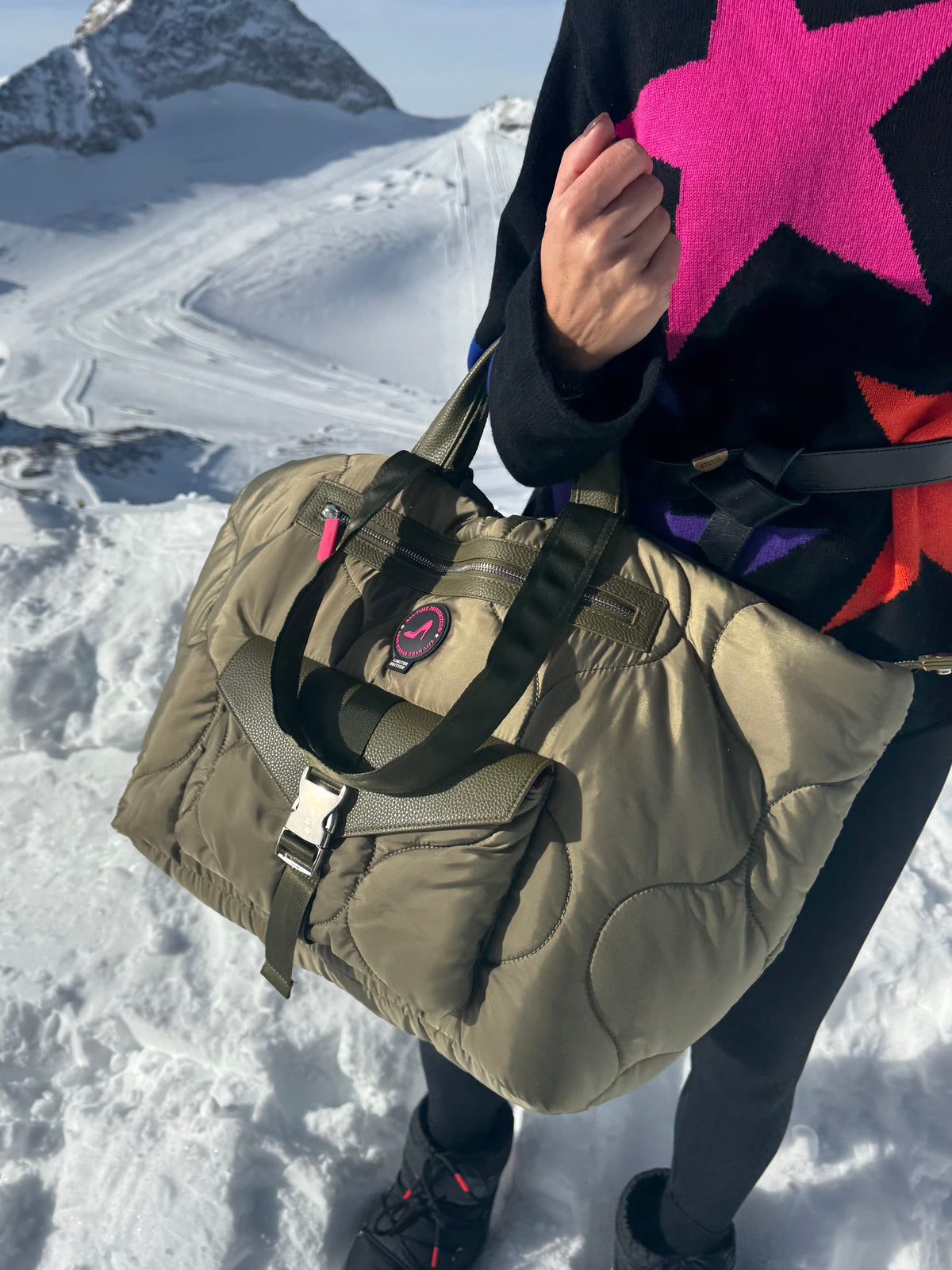 Person in snow holds puffy green medium tote bag, wearing black pants and a star-patterned sweater.