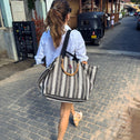 A woman with a ponytail walks down a sunlit street carrying the Medium Tote Bag Black & White GALLE with bamboo handles. She wears a light blue shirt, shorts, sandals, and sunglasses. A black tuk-tuk and people are in the background.