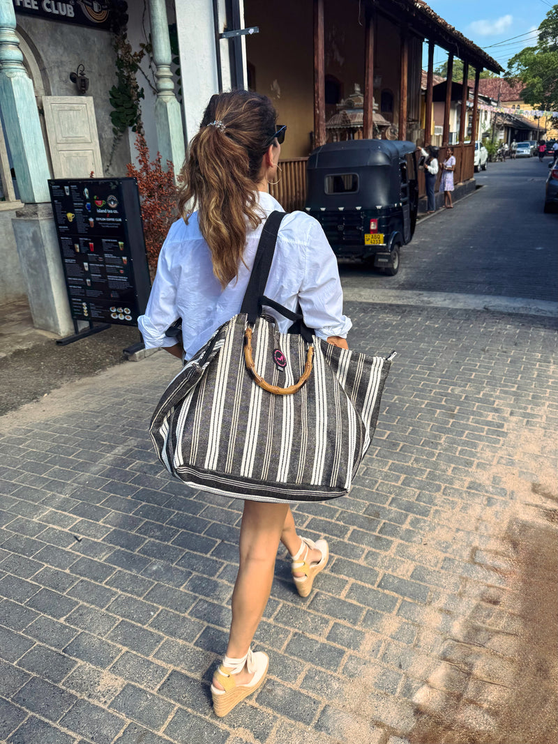 A woman with a ponytail walks down a sunlit street carrying the Medium Tote Bag Black & White GALLE with bamboo handles. She wears a light blue shirt, shorts, sandals, and sunglasses. A black tuk-tuk and people are in the background.