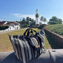The Medium Tote Bag Black & White GALLE with bamboo handles is displayed on a cannon, set against grass, palm trees, and a white lighthouse under a clear blue sky.