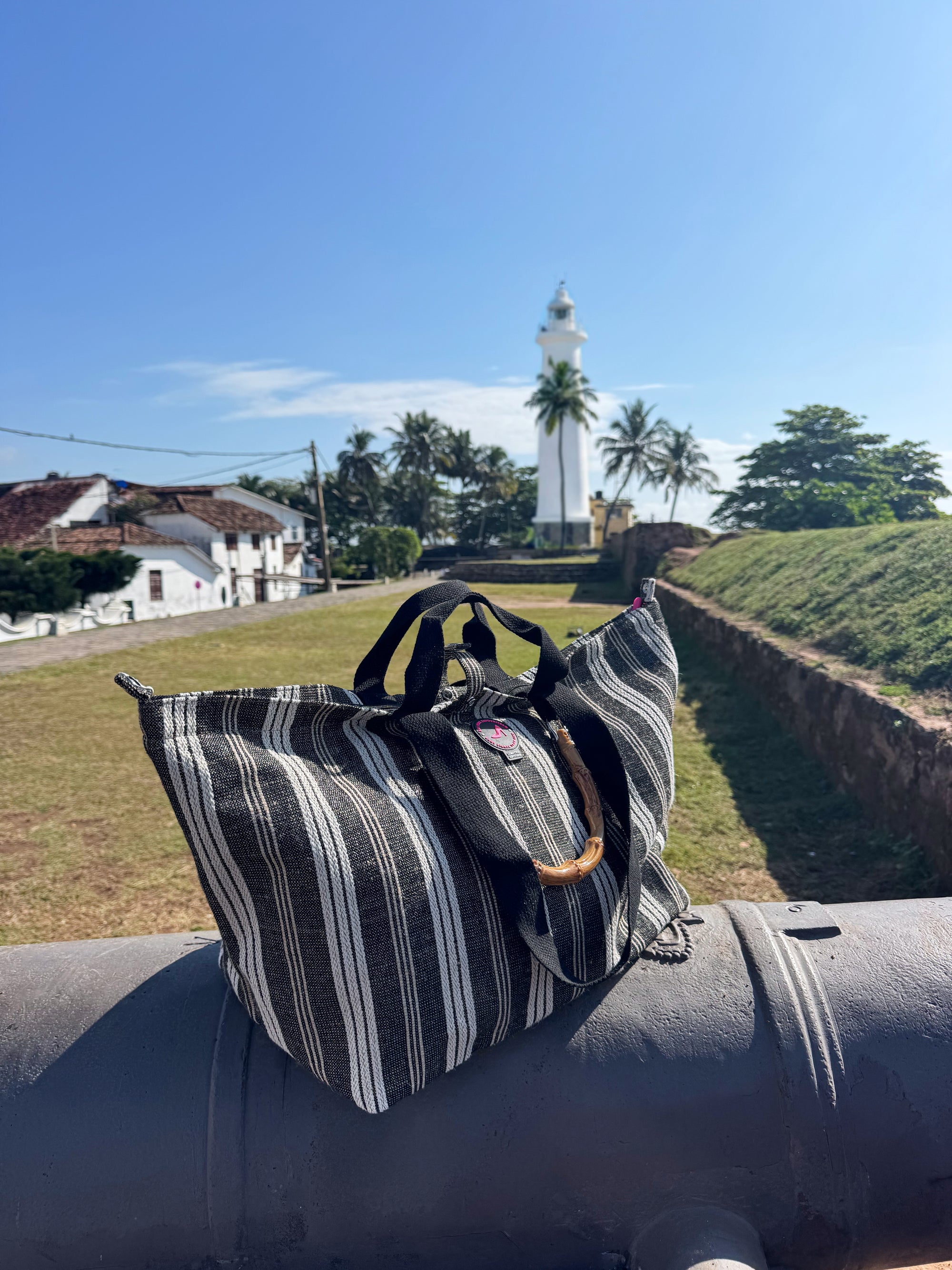 The Medium Tote Bag Black & White GALLE with bamboo handles is displayed on a cannon, set against grass, palm trees, and a white lighthouse under a clear blue sky.