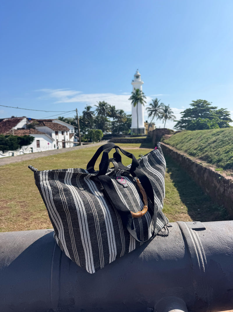 The Medium Tote Bag Black & White GALLE with bamboo handles is displayed on a cannon, set against grass, palm trees, and a white lighthouse under a clear blue sky.