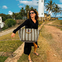 A woman in a black dress and sunglasses walks outdoors, smiling and carrying a Medium Tote Bag Black & White GALLE with bamboo handles. Palm trees, a stone pathway, and a tall white lighthouse rise behind her under a partly cloudy sky.