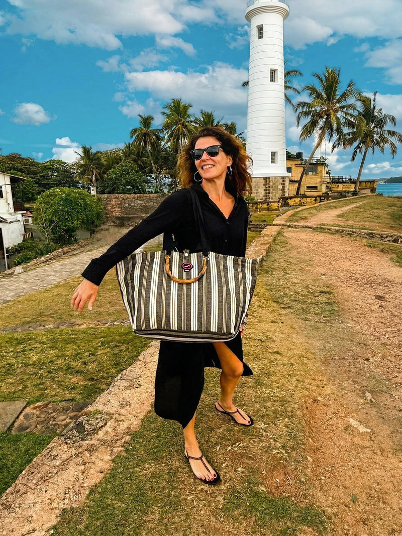 A woman in a black dress and sunglasses walks outdoors, smiling and carrying a Medium Tote Bag Black & White GALLE with bamboo handles. Palm trees, a stone pathway, and a tall white lighthouse rise behind her under a partly cloudy sky.