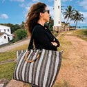 A woman in sunglasses and a black outfit stands outdoors near a white lighthouse, holding a Medium Tote Bag Black & White GALLE with bamboo handles. Palm trees and a partly cloudy sky are in the background.