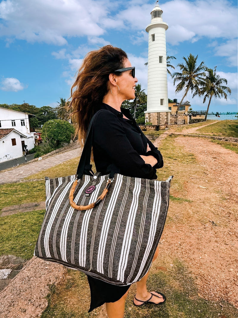 A woman in sunglasses and a black outfit stands outdoors near a white lighthouse, holding a Medium Tote Bag Black & White GALLE with bamboo handles. Palm trees and a partly cloudy sky are in the background.